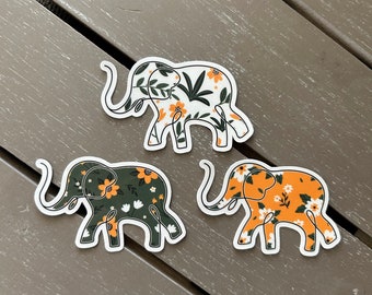 May include: Three elephant-shaped stickers featuring floral patterns. One sticker has a white background with orange flowers and green leaves. Another has a dark green background with orange and white flowers. The third has an orange background with white flowers.