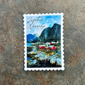Op de afbeelding: Een witte sticker in de vorm van een postzegel met een landschap van Lofoten, Noorwegen. De afbeelding toont rode gebouwen aan de kust, bergen en de tekst "Lofoten Norway". De sticker heeft een gekartelde rand.