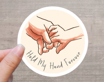 May include: A round sticker with a watercolor illustration of two hands holding each other. The text "Hold My Hand Forever" is written in a handwritten font around the bottom of the image. The sticker is held by a person's fingers.