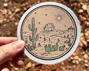 May include: A round sticker featuring a desert landscape with a saguaro cactus, prickly pear cacti, and a mesa under a sun and stars. The sticker has a beige background and a black border.