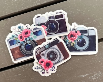 May include: Four stickers of vintage cameras with floral decorations. The cameras are in shades of brown, black, and white, each decorated with pink flowers and green foliage. The stickers are on a weathered, grey wooden surface.