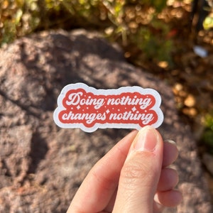May include: A white sticker with a red border and the text "Doing nothing changes nothing" in a cursive font. The sticker is held by a hand against a blurred natural background.