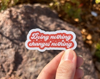 May include: A white sticker with a red border and the text "Doing nothing changes nothing" in a cursive font. The sticker is held by a hand against a blurred natural background.