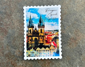 May include: A white, postage stamp-shaped magnet featuring a colorful image of Prague, Czech Republic. The image depicts a church with tall spires and red rooftops under a blue sky. The text "Prague, Czech Republic" is written in cursive.