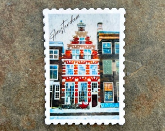 May include: A rectangular stamp featuring a colorful illustration of buildings in Amsterdam. The central building is red and white, with the word "Amsterdam" written in cursive at the top. The stamp has a white border.