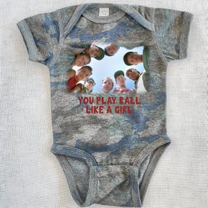 Peut inclure: Un body b&eacute;b&eacute; en camouflage gris et bleu avec une image d'un groupe de gar&ccedil;ons regardant la cam&eacute;ra. Le texte "YOU PLAY BALL LIKE A GIRL" est imprim&eacute; en rouge sur le body.