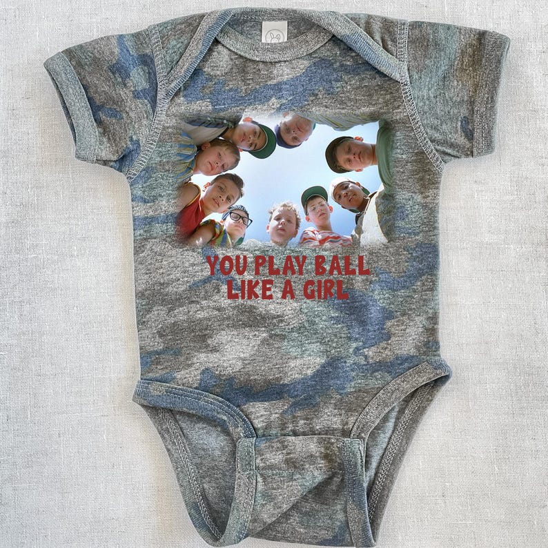 Peut inclure: Un body b&eacute;b&eacute; en camouflage gris et bleu avec une image d'un groupe de gar&ccedil;ons regardant la cam&eacute;ra. Le texte "YOU PLAY BALL LIKE A GIRL" est imprim&eacute; en rouge sur le body.
