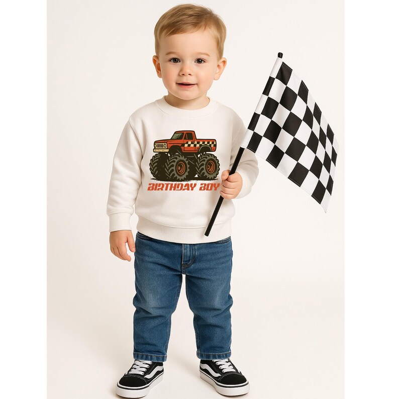 以下が含まれることがあります： 赤いモンスターカーのグラフィックと「BIRTHDAY BOY」の文字が入った白いスウェットシャツを着た幼い子供。子供はチェッカーフラッグを持ち、青いジーンズと白と黒のスニーカーを履いています。