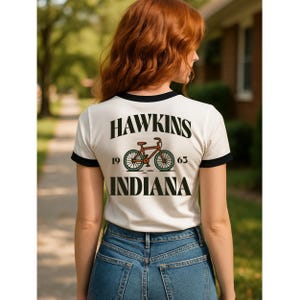 May include: White t-shirt with black trim featuring the text "HAWKINS INDIANA" and a bicycle graphic. The shirt includes the numbers "19" and "63" on either side of the bicycle. The person in the image is wearing blue jeans.