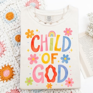 May include: Cream-colored t-shirt with the words "CHILD OF GOD" in colorful, whimsical lettering. The design includes a rainbow and flower graphics. A delicate necklace with butterfly charms rests on the shirt.