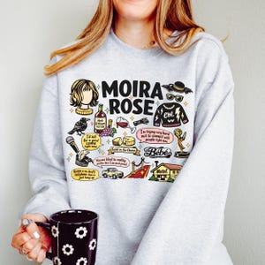 May include: A light gray sweatshirt featuring a graphic design with the words "MOIRA ROSE" and various illustrations. The design includes a wig, a crow, wine, cheese, and the text "ew."