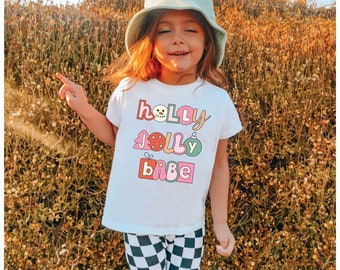 May include: A young child wearing a white t-shirt with the words "HOLLY JOLLY BABE" in colorful, retro-style lettering. The child is also wearing checkered pants and a light green bucket hat with a smiley face.