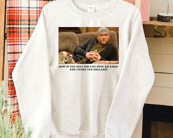 May include: A light gray sweatshirt featuring a rectangular image of a man on a couch. The image has the text: "HOW IN THE HELL DID YOU FIND AN X-BOX FOR UNDER TEN DOLLARS?"