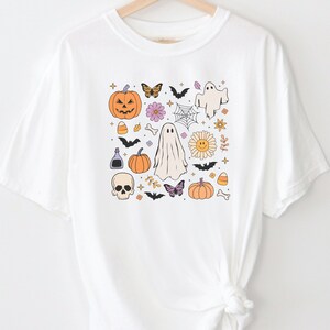 May include: A white short-sleeve t-shirt featuring a central graphic print of various Halloween-themed icons including orange pumpkins, white ghosts, black bats, a skull, a spider web, butterflies, flowers, candy corn, and small bones.