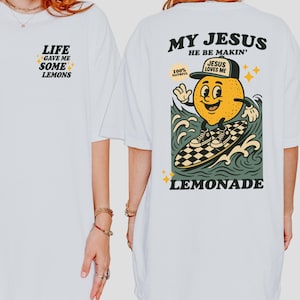 May include: White t-shirt with the text "LIFE GAVE ME SOME LEMONS" on the front. The back features a cartoon lemon surfing with the words "MY JESUS HE BE MAKIN'" and "LEMONADE". The lemon wears a hat that says "JESUS LOVES ME".