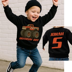 以下が含まれることがあります： 赤いモンスタートラックのグラフィックと「BIRTHDAY BOY」の文字が入った黒いスウェットシャツ。背面にはオレンジ色で「JONAH」という名前と数字の「5」が表示されています。男の子は黒いビーニーとブルージーンズを着用しています。