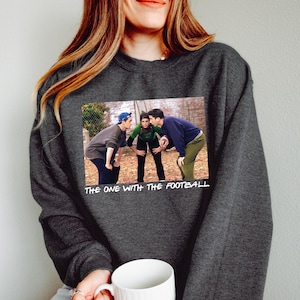 May include: Dark gray sweatshirt featuring a graphic of three people playing football, with the text "THE ONE WITH THE FOOTBALL" below the image. The sweatshirt is a solid color and has a crew neck.