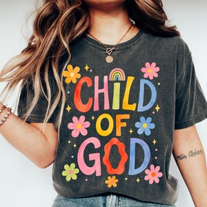 May include: Dark gray t-shirt with the words "CHILD OF GOD" in colorful, floral-themed lettering. The design includes a rainbow and flower graphics, creating a cheerful and inspirational aesthetic. The shirt is a casual wear item.