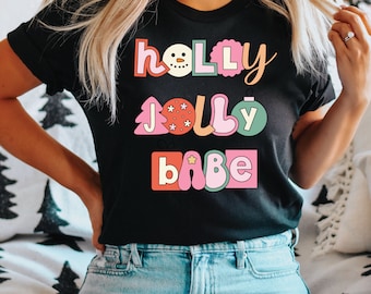 May include: Black t-shirt with the words "Holly Jolly Babe" in a retro font. The design includes a snowman, Christmas tree, and ornament. The letters are in pink, orange, and green.