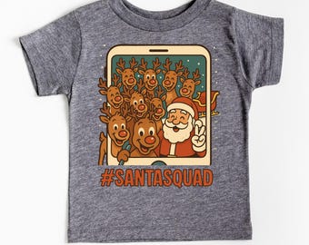May include: A gray toddler t-shirt featuring a cartoon image of Santa Claus and reindeer taking a selfie. The image is inside a phone-like frame, with the text "#SANTASQUAD" in orange.