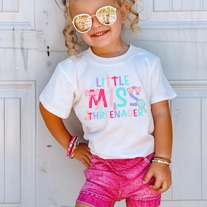 May include: A white t-shirt with the text "Little Miss Threenager" in colorful letters and graphics. The shirt features a pink bow, lipstick, and a magnifying glass.