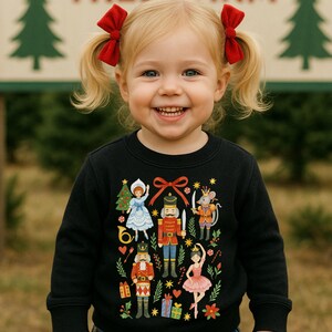 May include: A smiling child with blonde pigtails wearing a black sweatshirt. The sweatshirt features a colorful illustration of characters from The Nutcracker ballet, including a ballerina, a mouse, and a nutcracker soldier.