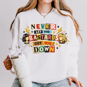May include: White sweatshirt with the text "NEVER LET THE BASTARDS GET YOU DOWN" in colorful, cut-out style letters. The design includes illustrations of women's faces and star and lightning bolt accents.