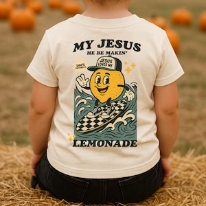 May include: Cream-colored t-shirt with a graphic of a lemon character surfing on a checkered surfboard. The shirt features the text "MY JESUS HE BE MAKIN'" and "LEMONADE". The lemon wears a hat that says "JESUS LOVES ME".
