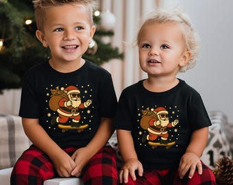 May include: Two children wearing matching black t-shirts with a Santa Claus graphic riding a skateboard. The Santa Claus illustration is in red, tan, and black. The children are also wearing red and black plaid pajama pants.