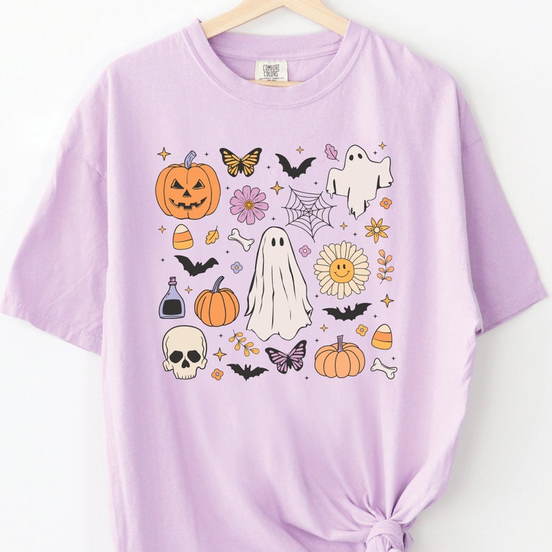 May include: A light purple short-sleeve t-shirt featuring a central graphic print of various Halloween-themed icons including orange pumpkins, white ghosts, a skull, bats, butterflies, a spider web, a smiley face flower, and candy corn.