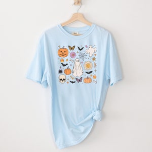 May include: A light blue t-shirt featuring a central graphic print of Halloween-themed icons including a jack-o'-lantern, a skull, ghosts, bats, butterflies, a spider web, a daisy with a smiley face, candy corn, and a potion bottle.