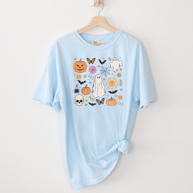May include: A light blue t-shirt featuring a central graphic print of Halloween-themed icons including a jack-o'-lantern, a skull, ghosts, bats, butterflies, a spider web, a daisy with a smiley face, candy corn, and a potion bottle.