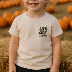 May include: A beige t-shirt with the text "LIFE GAVE ME SOME LEMONS" in black and yellow. The shirt is short-sleeved and appears to be made of cotton. The background shows pumpkins and hay bales.