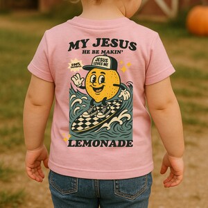 May include: Pink short-sleeved t-shirt with the text "MY JESUS HE BE MAKIN'" in black. The shirt features a cartoon lemon character surfing on a checkered surfboard, with the word "LEMONADE" below. The lemon wears a hat that says "JESUS LOVES ME".