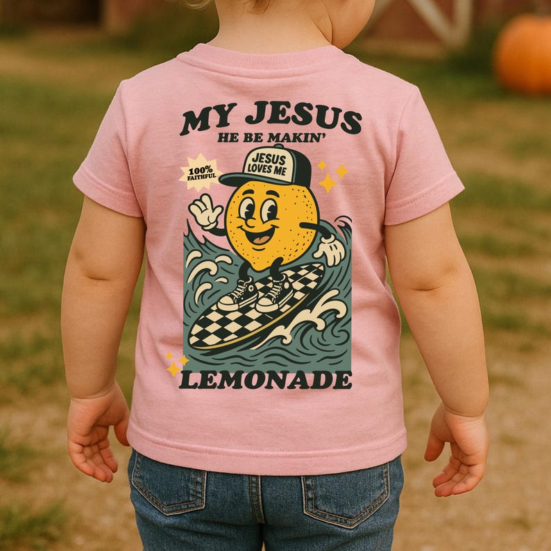 May include: Pink short-sleeved t-shirt with the text "MY JESUS HE BE MAKIN'" in black. The shirt features a cartoon lemon character surfing on a checkered surfboard, with the word "LEMONADE" below. The lemon wears a hat that says "JESUS LOVES ME".