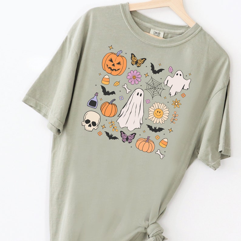 May include: A sage green short-sleeve t-shirt featuring a central graphic print of Halloween-themed icons including a jack-o'-lantern, ghosts, a skull, bats, butterflies, a spider web, a smiley face flower, candy corn, and a potion bottle.