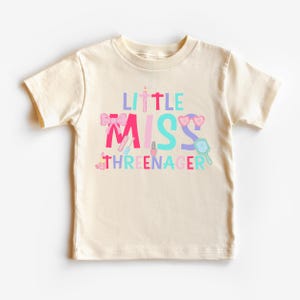 May include: A cream colored t-shirt with the text "Little Miss Threenager" in pink, blue, and purple. The text is surrounded by images of makeup, a bow, and sunglasses.