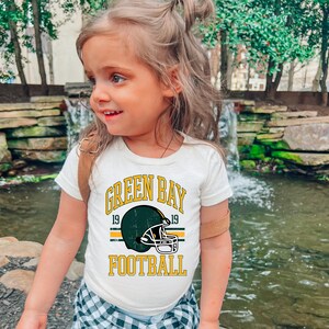 Puede incluir: Camiseta color crema con un gr&aacute;fico de casco de f&uacute;tbol americano de los Green Bay Packers. El dise&ntilde;o incluye las palabras "GREEN BAY FOOTBALL" en amarillo, con el a&ntilde;o "19" a cada lado del casco. La camiseta la lleva un ni&ntilde;o peque&ntilde;o.