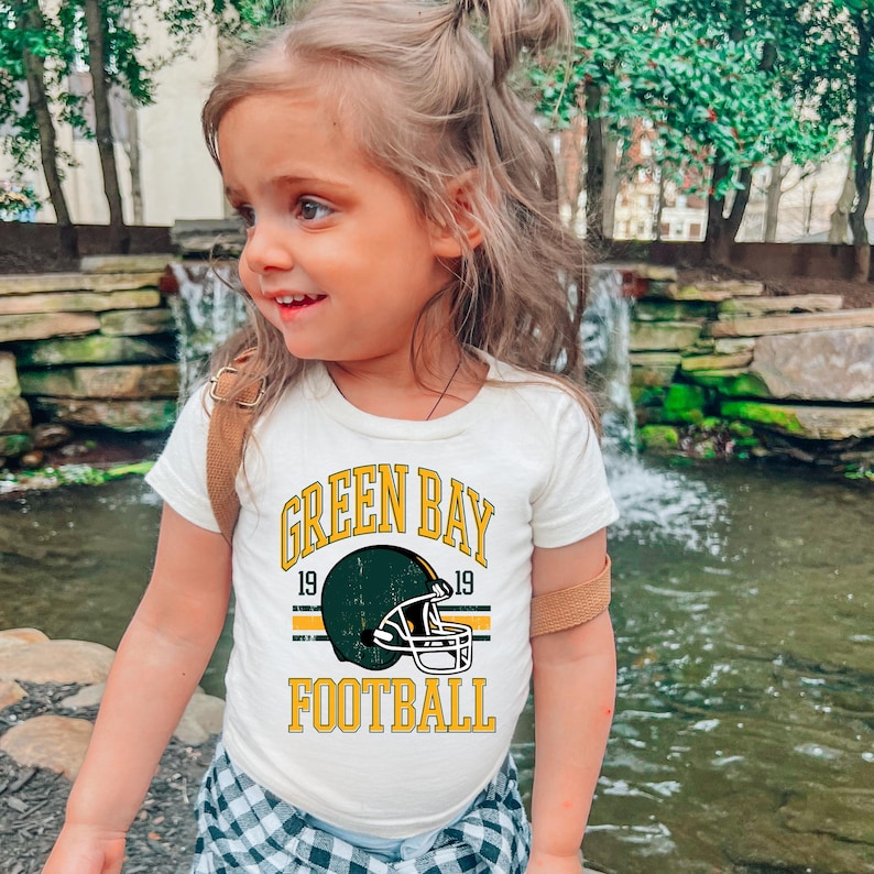 Puede incluir: Camiseta color crema con un gr&aacute;fico de casco de f&uacute;tbol americano de los Green Bay Packers. El dise&ntilde;o incluye las palabras "GREEN BAY FOOTBALL" en amarillo, con el a&ntilde;o "19" a cada lado del casco. La camiseta la lleva un ni&ntilde;o peque&ntilde;o.