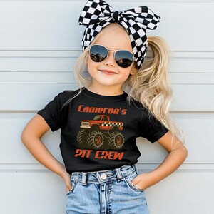 Pu&ograve; includere: Una maglietta nera con la scritta "Cameron's PIT CREW" in arancione e una grafica di un monster truck rosso. Il bambino indossa occhiali da sole e un fiocco a scacchi bianco e nero.