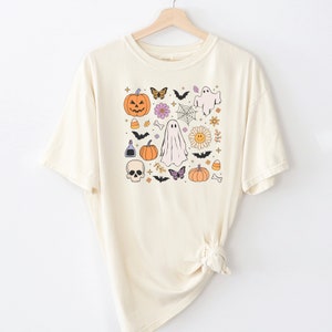 May include: A cream-colored short-sleeve t-shirt featuring a central graphic print of various Halloween-themed icons including pumpkins, ghosts, a skull, bats, butterflies, flowers, a spider web, candy corn, and bones arranged in a square pattern.