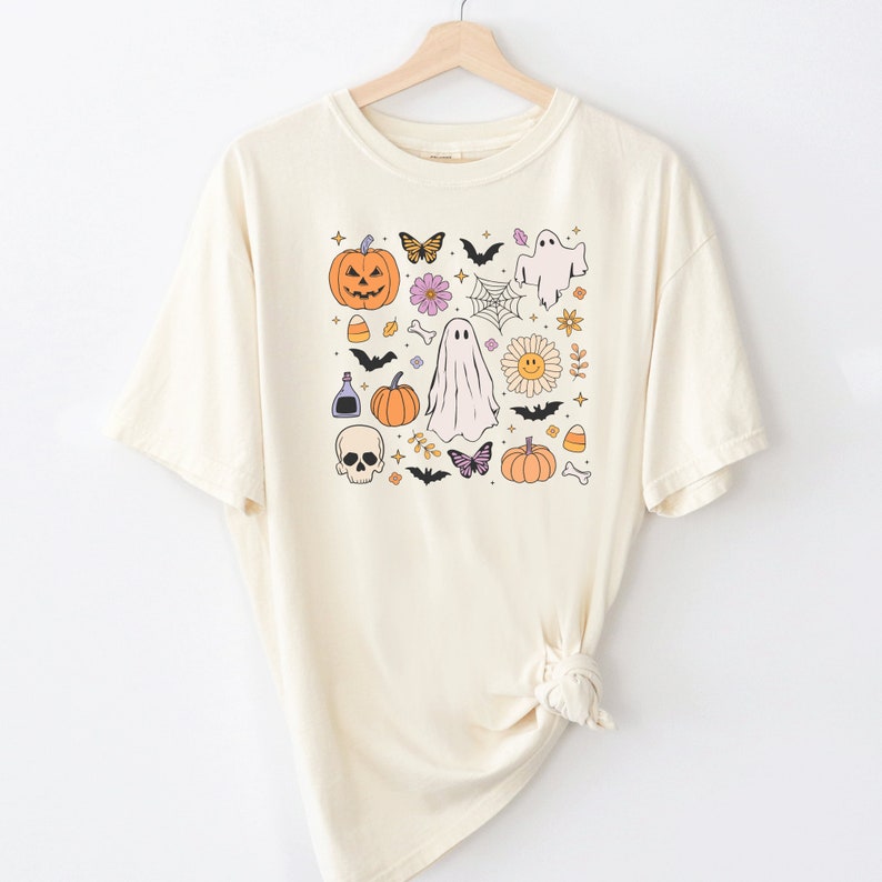 May include: A cream-colored short-sleeve t-shirt featuring a central graphic print of various Halloween-themed icons including pumpkins, ghosts, a skull, bats, butterflies, flowers, a spider web, candy corn, and bones arranged in a square pattern.