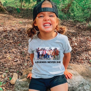 May include: A young person wearing a grey t-shirt with a black and white image of a group of people and the text "Legends Never Die". The person is wearing a black baseball cap and has a happy expression.