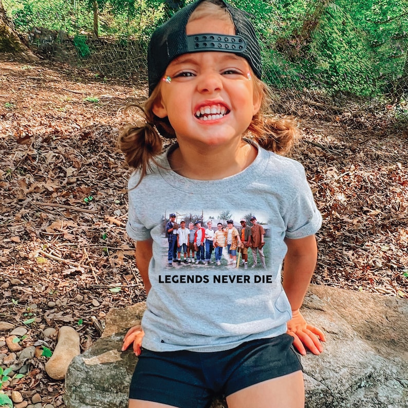May include: A young person wearing a grey t-shirt with a black and white image of a group of people and the text "Legends Never Die". The person is wearing a black baseball cap and has a happy expression.