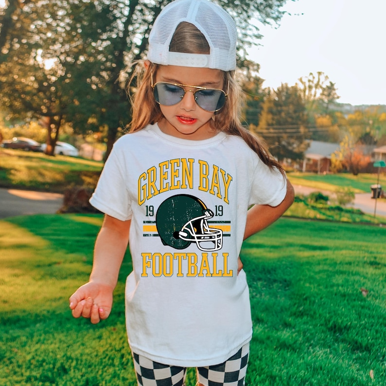 Puede incluir: Camiseta blanca con un dise&ntilde;o de f&uacute;tbol de los Green Bay Packers. La camiseta presenta las palabras "GREEN BAY FOOTBALL" en amarillo, con un gr&aacute;fico de casco de f&uacute;tbol verde y amarillo. La camiseta la lleva una persona con una gorra blanca y gafas de sol.