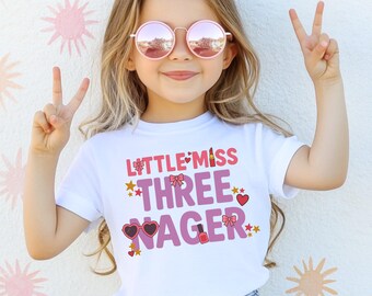 May include: A white t-shirt with the text "LITTLE MISS THREE NAGER" in pink and purple, decorated with stars, hearts, and makeup-themed graphics. The girl is wearing pink sunglasses and making a peace sign.