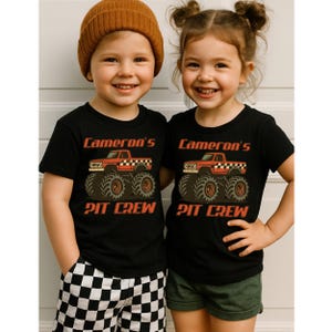 Pu&ograve; includere: Due bambini con magliette nere con una grafica di monster truck rosso e la scritta "Cameron's Pit Crew". Uno indossa un berretto marrone e pantaloncini a scacchi, l'altro pantaloncini verdi.