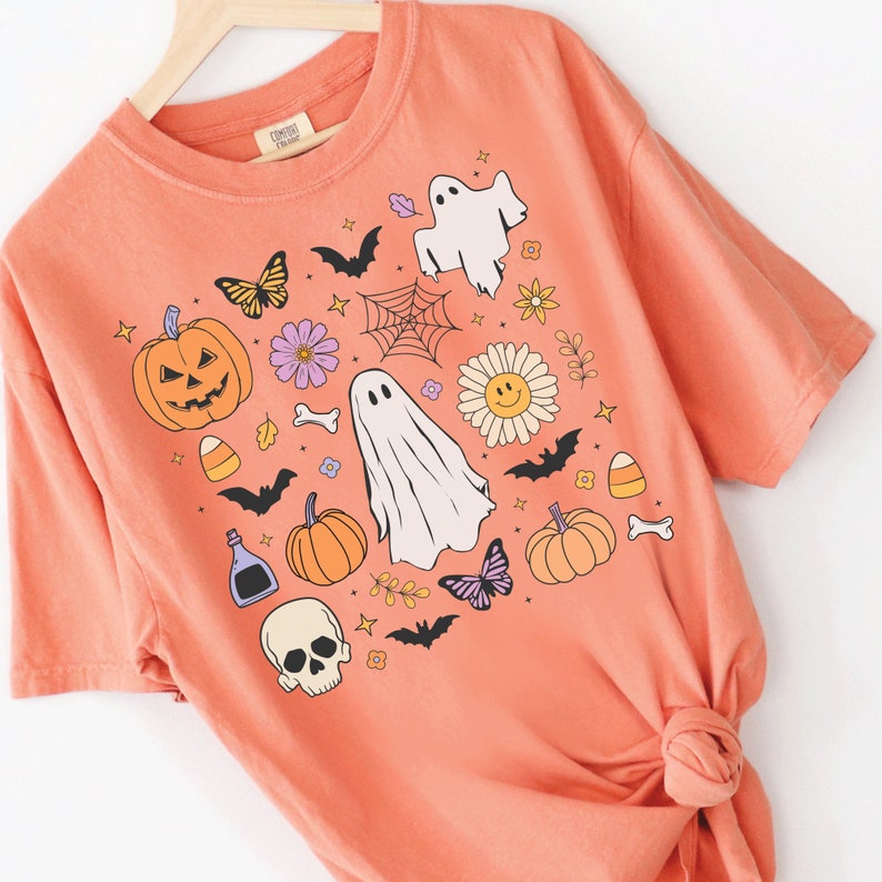 May include: A coral-colored short-sleeve t-shirt featuring a graphic print of Halloween-themed icons including ghosts, jack-o'-lanterns, skulls, bats, spiders, candy corn, flowers, and butterflies arranged in a central cluster on the front.