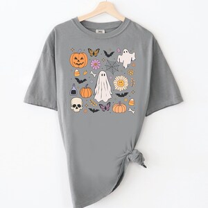 May include: A grey short-sleeve t-shirt featuring a graphic print of Halloween-themed icons including a jack-o'-lantern, ghosts, a skull, bats, butterflies, a spider web, a smiley face flower, candy corn, a potion bottle, and small bones.