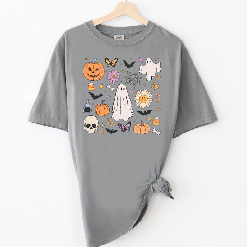 May include: A grey short-sleeve t-shirt featuring a graphic print of Halloween-themed icons including a jack-o'-lantern, ghosts, a skull, bats, butterflies, a spider web, a smiley face flower, candy corn, a potion bottle, and small bones.